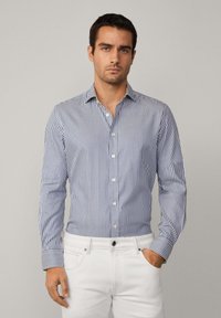 Striped button-up shirt in blue and white, long sleeves, slim fit, subtle texture, paired with white jeans, featuring a classic collar.