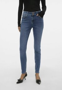 Jeans skinny azul con cierre de botón, cinco bolsillos y ligeros detalles de desgaste, combinados con zapatos de tacón negros.