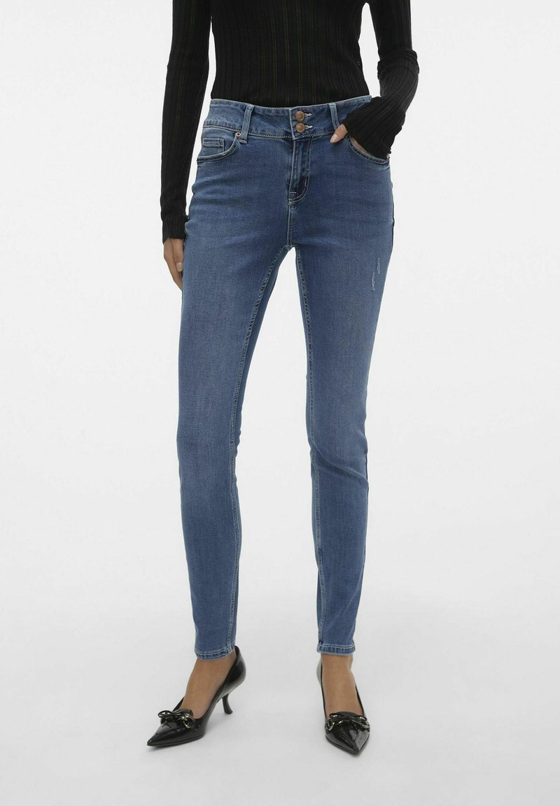 Jeans skinny azul con cierre de botón, cinco bolsillos y ligeros detalles de desgaste, combinados con zapatos de tacón negros.
