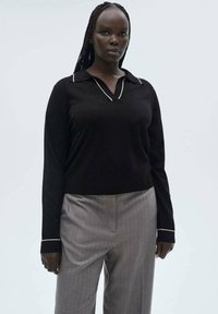 Schwarzer Strickpullover mit Polo-Kragen und weißen Akzenten, kombiniert mit hellgrauen Hosen, die eine glatte Textur und einen lässigen Schnitt aufweisen.