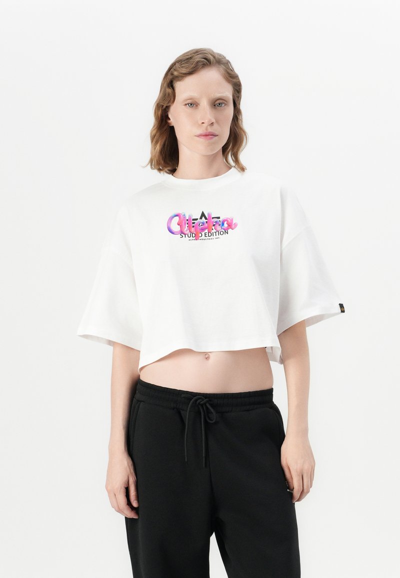 Joven con cabello corto y ondulado, vistiendo una camiseta blanca ajustada con texto colorido y pantalones negros con cordón, de pie contra un fondo sencillo.
