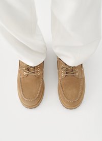 Καφέ παπούτσια σουέντ με σχέδιο μοκασίνι, με κλείσιμο με κορδόνια και αντίθετη ραφή, σε συνδυασμό με λευκό παντελόνι.