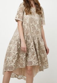 Femme portant une robe en dentelle beige à manches courtes, motifs floraux, et ourlet asymétrique, debout devant un fond uni.