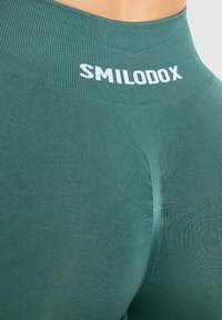 Leggings verdi senza cuciture con una texture a coste, con un logo bianco "SMILODOX" nella vita e un design liscio e aderente.