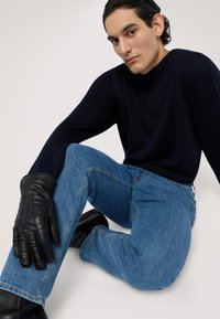Czarne skórzane rękawiczki kontrastują z granatowym swetrem i jasnoniebieskimi dżinsami. Model siedzi, prezentując zrelaksowaną postawę i faktury.