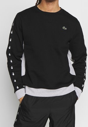 Man draagt een zwart-grijs sweatshirt met lange mouwen, met witte krokodillenlogo's langs de mouwen en een klein groen krokodillenlogo op de borst.