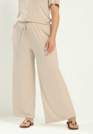 Person iført beige bukser med vide ben og snøre, beige kortærmet top og brune sandaler med gyldne detaljer, stående med den ene hånd i lommen.