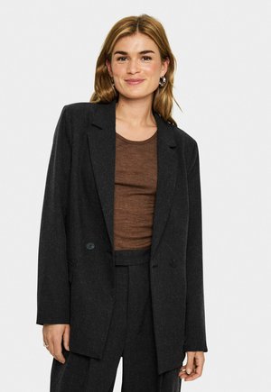 Donna con capelli biondi alla spalla indossa un grande blazer nero e un top marrone, sorridendo dolcemente su uno sfondo chiaro e semplice.