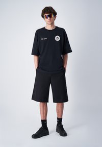 Zwarte oversized t-shirt met logo, gecombineerd met zwarte shorts. Donkere sneakers en patroon zonnebril maken de outfit compleet. Eenvoudig, casual ontwerp.
