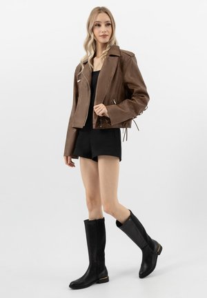 Junge Frau in einer braunen Lederjacke, schwarzen Shorts und schwarzen kniehohen Stiefeln, posiert vor einem schlichten, hellen Hintergrund.