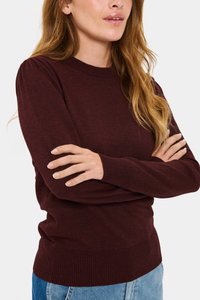 Bordowy sweter z dzianiny z długimi rękawami, żebrowanym dekoltem i dołem, miękka tekstura, noszony z rękami skrzyżowanymi na jasnoniebieskich dżinsach.