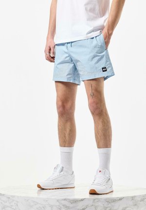 Lys blå shorts laget av et glatt stoff, med elastisk midje og sidelommer. Kombinert med hvite joggesko og hvite sokker.