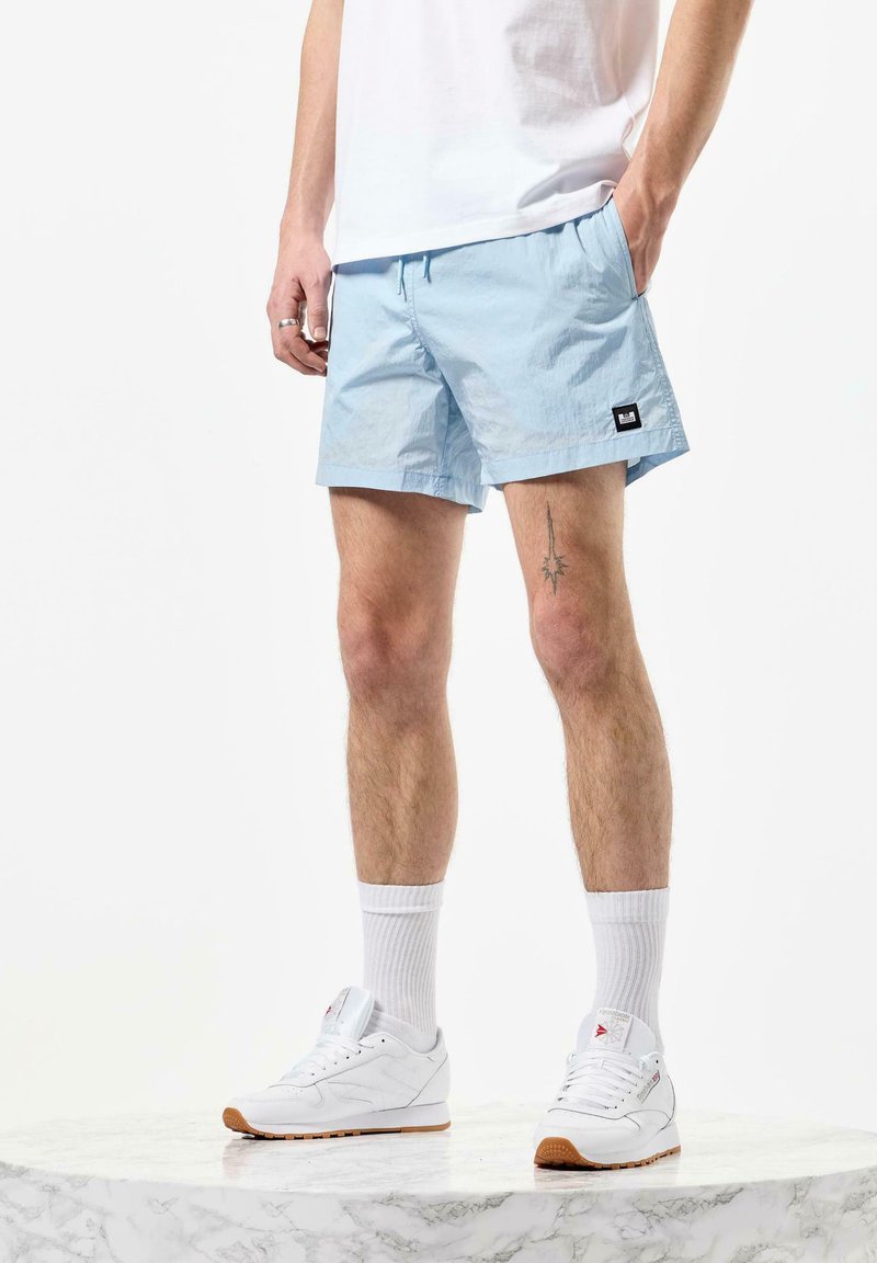 Shorts bleu clair en tissu lisse, avec une taille élastique et des poches latérales. Associés à des baskets blanches et des chaussettes blanches.