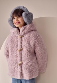 Cappotto rosa chiaro soffice con chiusure a bottoni in legno e ampio cappuccio, abbinato a paraorecchie grigi pelosi, indossato sopra jeans blu.