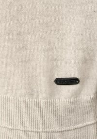 Ljusbeige stickad tröja med ribbade muddar och nederkant, med en liten svart etikett sydd i tyget. Mjuk textur och enkel design.