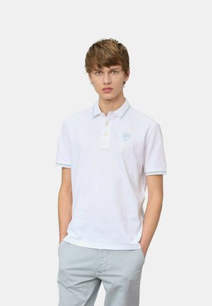 Giovane uomo con capelli castano chiaro che indossa una polo bianca a maniche corte e pantaloni grigio chiaro con le mani in tasca su sfondo bianco.