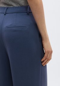 Σκούρο μπλε παντελόνι σε στενή γραμμή με ομαλή υφή, που διαθέτει πίσω τσέπη κ αι θηλιές για ζώνη, εμφανίζεται από το πλάι με το χέρι να ακουμπάει στη μέση του παντελονιού.