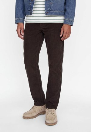 Homme portant un pantalon marron foncé, des chaussures beiges à lacets, une chemise rayée et une veste en jean, debout devant un fond clair uni.