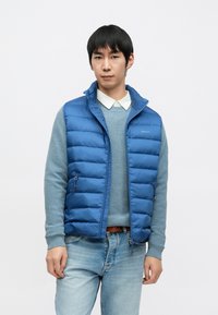 Vestă albastră tip puf cu fermoar frontal, asortată cu un pulover albastru deschis și blugi decolorati. Are un guler în picioare și buzunare cu fermoar.