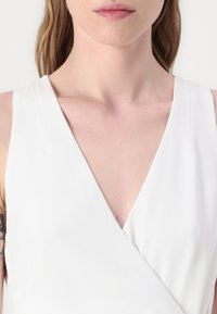 Top alb fără mâneci, cu un decolteu adânc în V și un design înfășurat. Material neted cu o textură subtilă, care evidențiază decolteul și umerii.