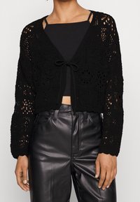 Černý háčkovaný kardigan s otevřenými vzory, zavazováním vpředu a dlouhými rukávy. Spárovaný s černým topem a koženými kalhotami.