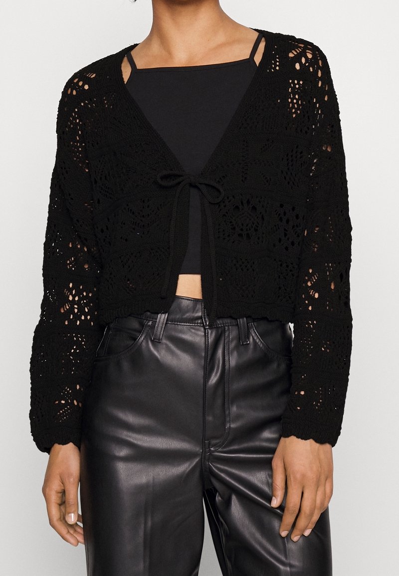 Černý háčkovaný kardigan s otevřenými vzory, zavazováním vpředu a dlouhými rukávy. Spárovaný s černým topem a koženými kalhotami.