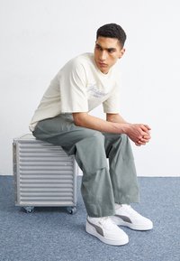 Ljusbeige bomulls t-shirt med blå text, ljusgröna cargobyxor, och vita sneakers med grå accenter, sittande på en silverförvaringslåda.