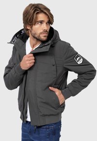 Gray zipped jacket with a hood, featuring a patch on the left arm, two front pockets, and a soft inner lining with a patterned design.