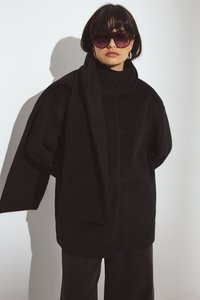 Czarny oversize płaszcz z wysokim kołnierzem i długim detalem szalowym, wykonany z miękkiego, teksturowanego materiału. Noszony z ciemnymi spodniami i dużymi okularami przeciwsłonecznymi.