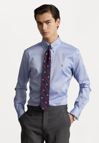 Chemise bleu clair à col boutonné, associée à une cravate marine avec des rayures diagonales rouges et des motifs brodés. Pantalons gris.