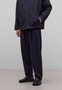 Camisa de lino azul marino con corte recto, dos bolsillos en el pecho y pantalones de ajuste relajado combinados con zapatos negros sin cordones. Textura suave y diseño minimalista.