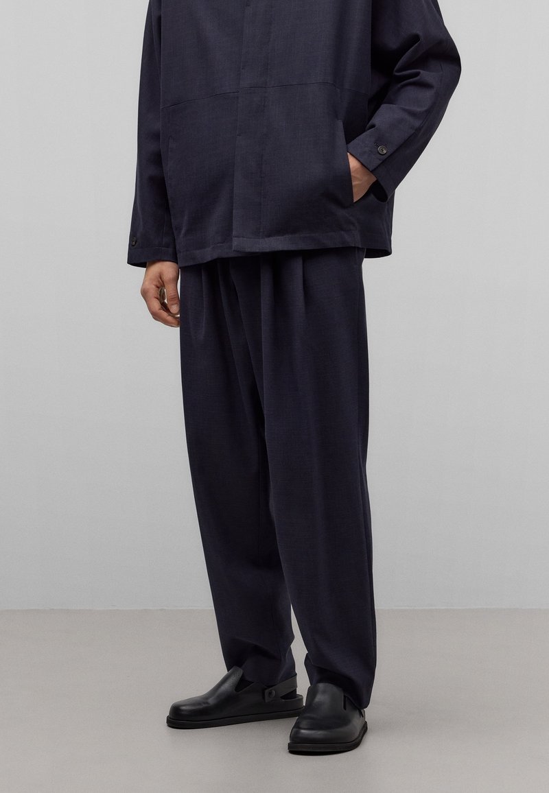 Camisa de lino azul marino con corte recto, dos bolsillos en el pecho y pantalones de ajuste relajado combinados con zapatos negros sin cordones. Textura suave y diseño minimalista.