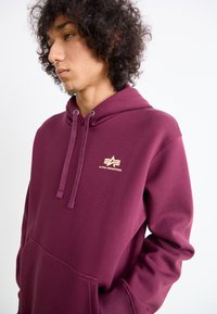 Joven con cabello rizado que lleva una sudadera de Alpha Industries en color burdeos con bolsillo delantero y cordones, mirando ligeramente de lado.