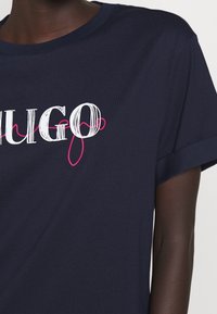 Granatowa bawełniana koszulka z krótkim rękawem z grafiką tekstową w kolorach białym i różowym "HUGO" na przodzie.