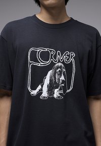 Μαύρο βαμβακερό μπλουζάκι με λευκό γραφικό σχέδιο ενός μπασέ χάουντ και στιλιζαρισμένο κείμενο "CORNER" μέσα σε ορθογώνιο πλαίσιο.