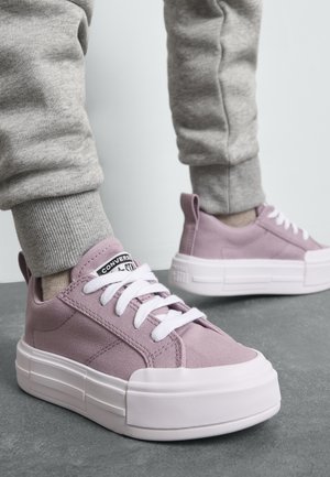 Roza platnena športna obutev z belimi vezalkami, debelimi belimi gumijastimi podplati in oblazinjeno ovratnikom leži na sivi površini, v kombinaciji s sivimi trenirkami.