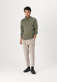 Olivgrüner Pullover mit Reißverschluss am Hals, langen Ärmeln sowie gerippten Bündchen und Saum. Kombiniert mit hellbeigen, schmal geschnittenen Hosen und schwarzen Slippers.