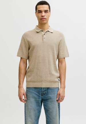 Mann iført beige kortermet strikket poloshirt og blå jeans, stående mot en enkel lys bakgrunn, vendt fremover.