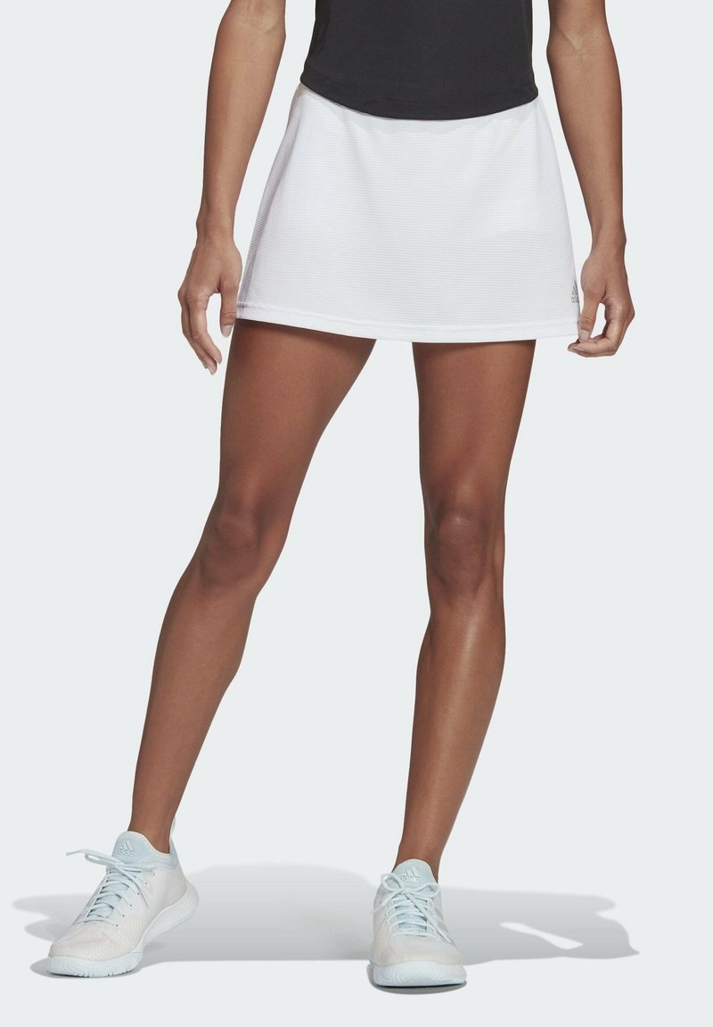 Falda atlética blanca con dobladillo recto, hecha de tejido texturizado, combinada con zapatillas de deporte azul claras. La parte superior es negra y ajustada.
