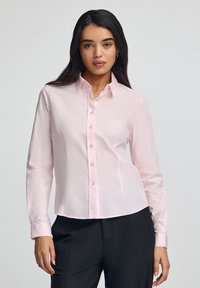 Camicia button-up rosa chiaro con colletto, maniche lunghe e leggera lucentezza. Presenta bottoni in tessuto e un logo ricamato sul lato sinistro del petto.