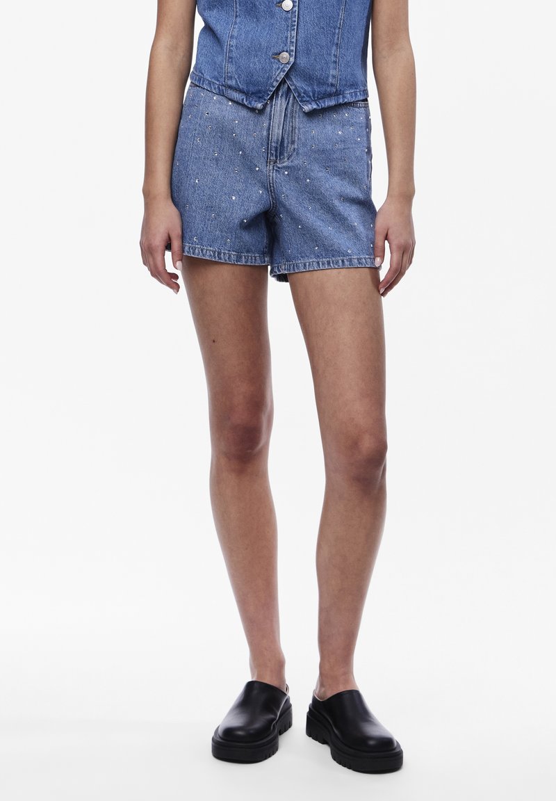 Short en denim orné de petites perles blanches, porté avec un gilet en denim assorti à boutons et des chaussures plates noires à plateforme sans lacets.