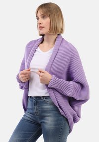 Lilla kardigan ribbknitiga, avtäckt fram och avslappnade ärmar, lager på valge valge T-särk, koos siniste teksadega.