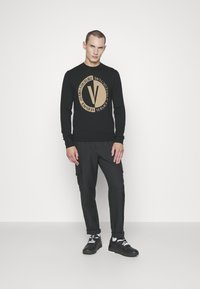 Zwarte trui met een gouden rond logo en tekst. Gecombineerd met zwarte cargo-broeken en sneakers, voorzien van witte veters en een gestructureerde zool.