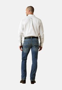 Camisa blanca de manga larga con puños abotonados, combinada con vaqueros azules desgastados. Cuenta con dos bolsillos traseros y una silueta ajustada.