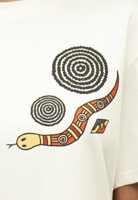 Στιλιζαρισμένο κόκκινο και μπεζ φίδι με γεωμετρικά μοτίβα και δύο ομόκεντρες μαύρες κυκλικές σχεδιάσεις σε λευκό ύφασμα.