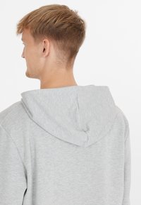 Ung man med kort ljusbrunt hår sedd bakifrån, iklädd en ljusgrå huvtröja mot en enkel vit bakgrund.