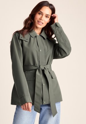 Young woman with long brown hair wearing an olive green belted jacket and light blue jeans, touching her hair with one hand.