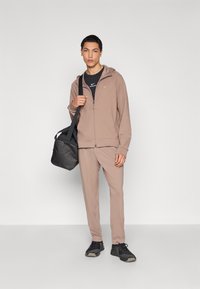 Ανοιχτό καφέ φούτερ με φερμουάρ, ταιριαστά παντελόνια φόρμας και μαύρα αθλητικά παπούτσια, με μαύρη τσάντα. Διαθέτει λογότυπο Nike στο στήθος και στο φούτερ.