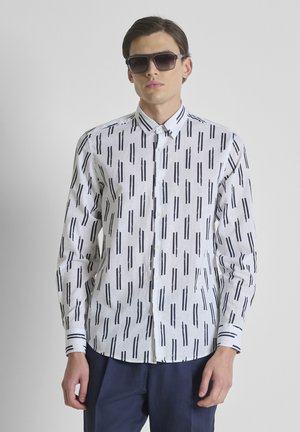 Homme portant des lunettes de soleil foncées, une chemise blanche à rayures noires verticales boutonnée et un pantalon bleu foncé, debout devant un fond uni.