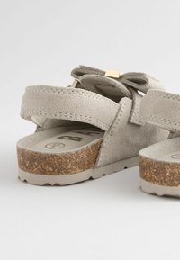 Beżowe zamszowe sandały na korkowej podeszwie, z regulowanymi paskami na kostce i dekoracyjnymi kokardkami na białym tle.